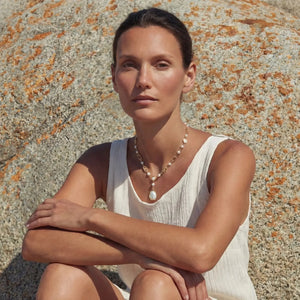 Woman sitting against a textured stone wall wearing a white sleeveless top and necklace.
