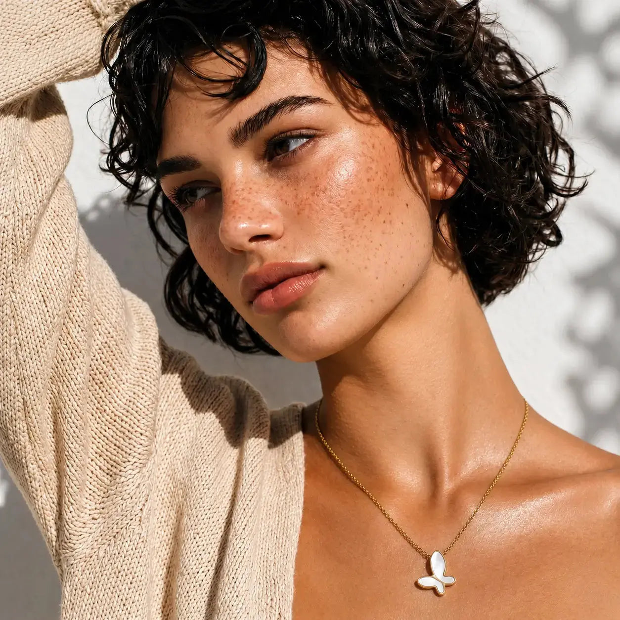 A close-up lifestyle shot of a woman with short curly dark hair wearing a delicate gold chain necklace with a white mother-of-pearl butterfly pendant. She is wearing a beige knit cardigan against a sunlit, textured white wall.