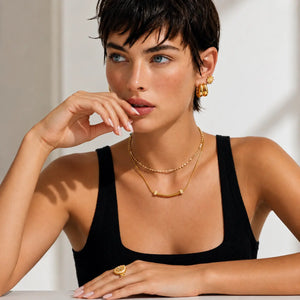 A woman with short dark hair wearing layered gold necklaces, hoop earrings, and a ring, posing in a black ribbed tank top against a bright white background.