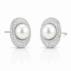 A pair of oval stud earrings featuring a central white pearl surrounded by a halo of small clear pavé stones in a silver-toned setting, shown on a clean white background with a soft reflection below.