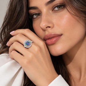 A close-up lifestyle shot of a woman wearing a silver-toned halo ring with a round center stone featuring blue and purple hues, set against a soft neutral background.