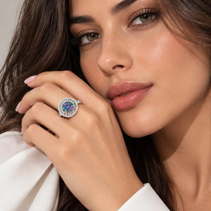 A close-up lifestyle shot of a woman wearing a silver-toned halo ring with a round center stone featuring blue and purple hues, set against a soft neutral background.