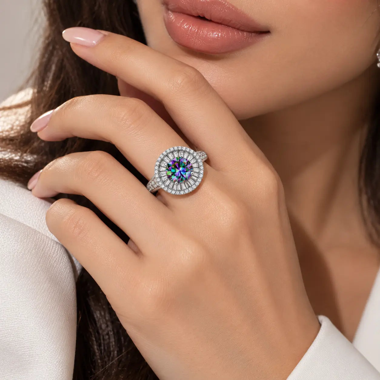 A close-up lifestyle shot of a model's hand wearing a silver-toned ring with a round iridescent blue gemstone surrounded by a halo of small clear stones.