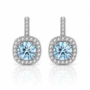 A pair of silver-toned halo stud earrings featuring a round light blue center stone surrounded by a halo of small clear stones, set against a white background with a soft reflection below.