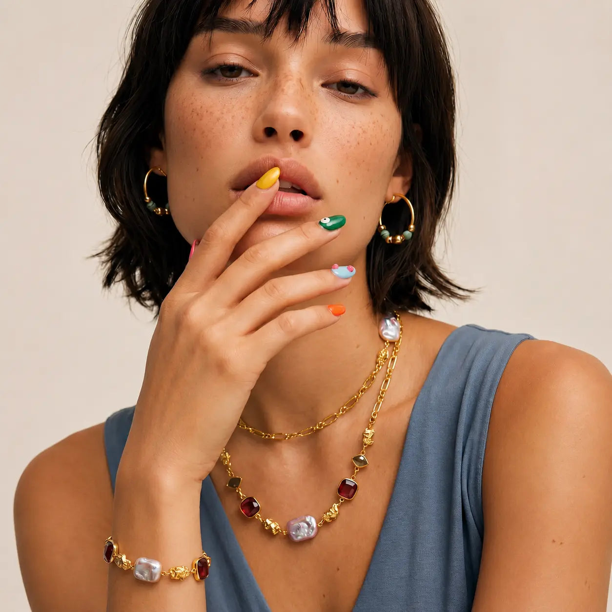 A close-up lifestyle shot of a model wearing a gold-toned jewelry set featuring a multi-colored stone necklace, matching bracelet, and hoop earrings. The model has short dark hair and is wearing a slate blue top against a plain beige background.