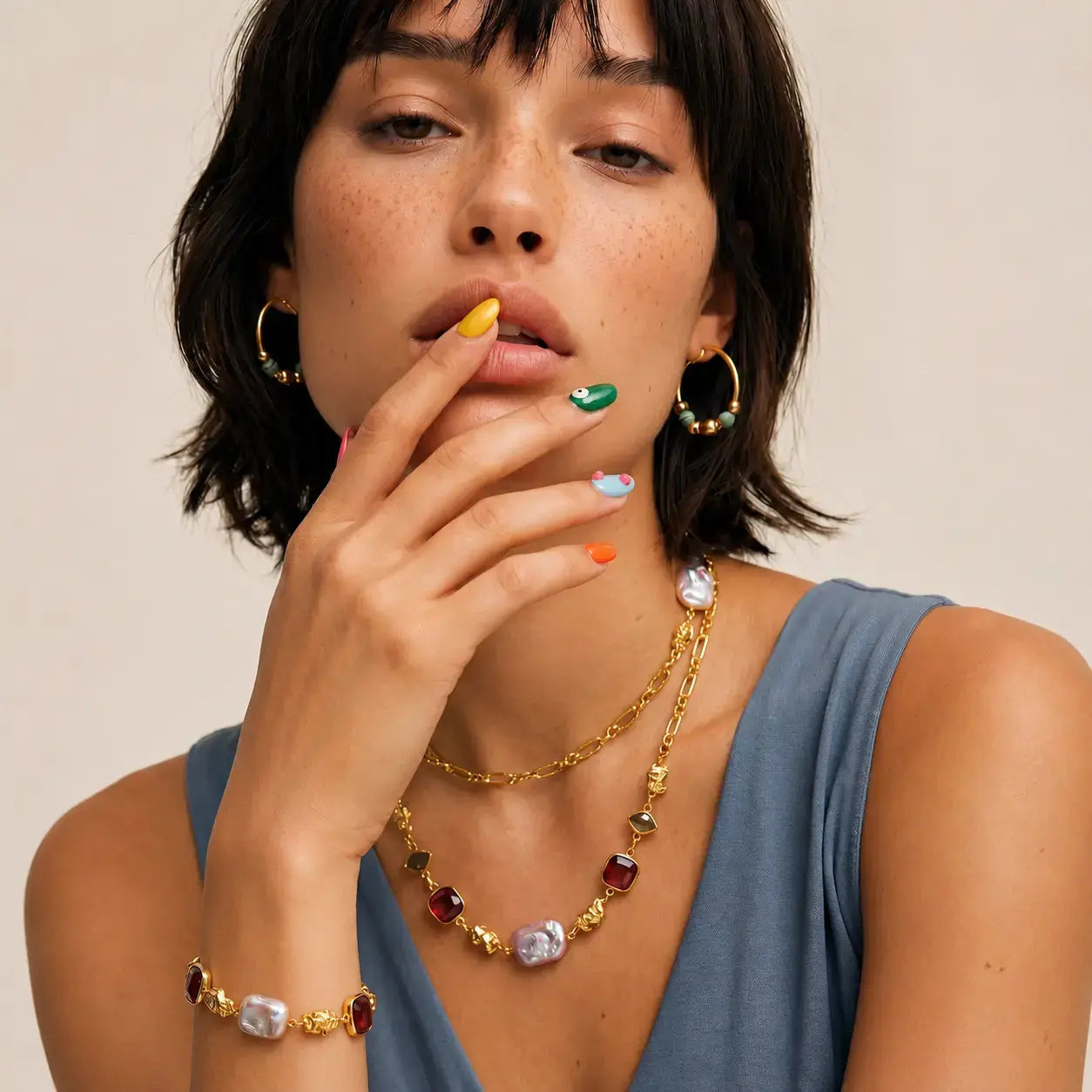 A close-up lifestyle shot of a model wearing a gold-toned jewelry set featuring a multi-colored stone necklace, matching bracelet, and hoop earrings. The model has short dark hair and is wearing a slate blue top against a plain beige background.