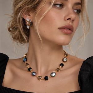 A close-up of a woman wearing a gold-toned necklace with oval iridescent shell-like stones and matching stud earrings against a soft beige background.
