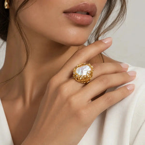 A close-up lifestyle shot of a model wearing a gold ring with a large, textured white mother-of-pearl cabochon on her finger.