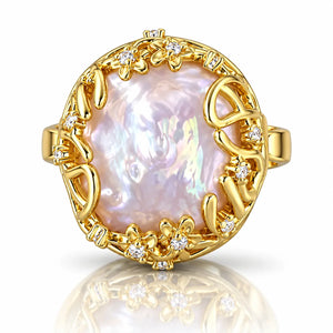 A gold ring featuring a large, round iridescent mother-of-pearl center stone surrounded by delicate floral gold settings and small clear accent stones, shown on a white background with a soft reflection below.
