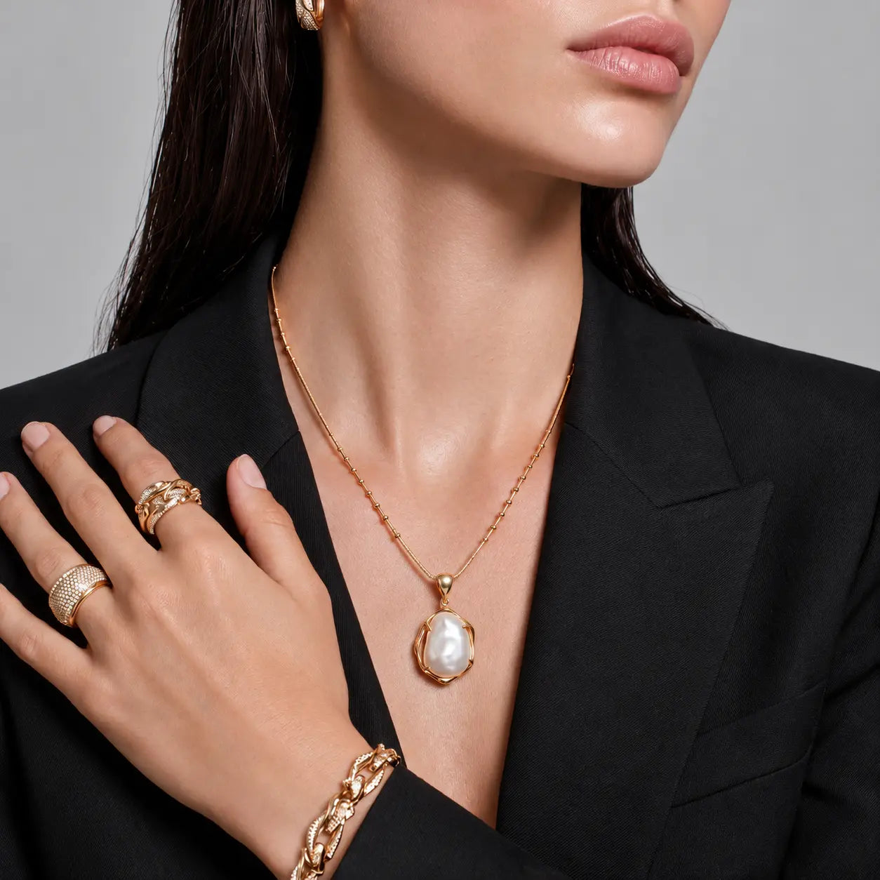 A close-up of a model wearing a gold pendant necklace with a large white pearl, along with matching gold rings and a bracelet, styled with a black blazer.
