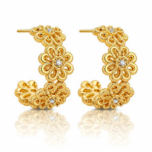 A pair of gold huggie hoop earrings with intricate floral filigree detailing and small clear crystal accents, displayed on a clean white background with a soft reflection below.