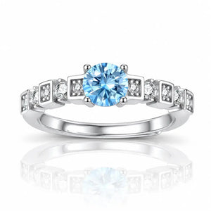 A silver-toned diamond ring featuring a round-cut light blue center stone and small clear accent stones set in square-shaped bands, shown on a white background with a soft reflection below.
