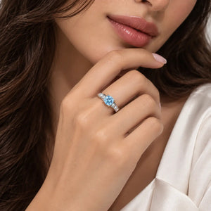 A close-up lifestyle shot of a woman wearing a silver ring with a central light blue gemstone and small side stones, resting her chin on her hand.