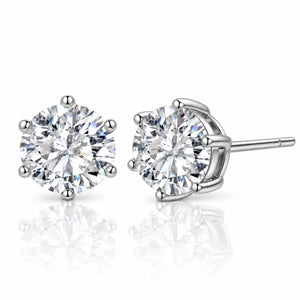 A pair of round-cut diamond stud earrings in a silver four-prong setting, with one earring facing forward and the other angled to show the post and back, on a reflective white surface.