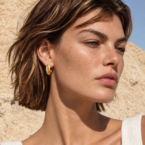 A close-up of a woman with short brown hair wearing a small gold crescent-shaped hoop earring, set against a textured beige stone wall and blue sky.