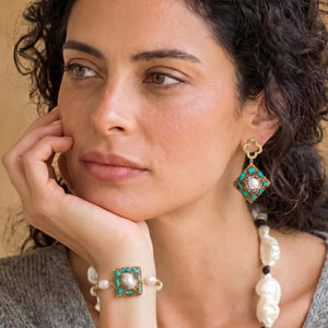 Woman wearing gold and turquoise earrings and bracelet, close-up on face and hand.
