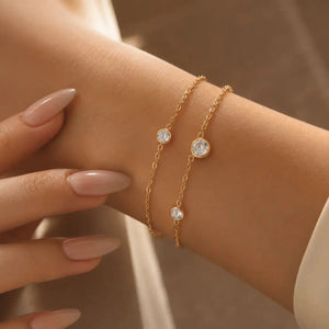 Two delicate gold chain bracelets with round bezel-set clear stones, shown worn on a woman's wrist with her fingers nearby.