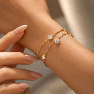 A close-up of a person's wrist wearing two delicate gold chain bracelets, each adorned with a round clear gemstone in a bezel setting. The person's hand and wrist are shown against a softly blurred neutral background.