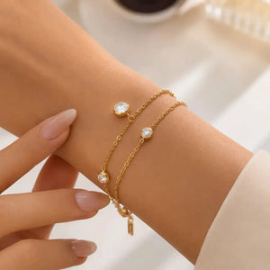 A close-up of a person's wrist wearing two delicate gold chain bracelets, each adorned with a round clear stone, against a softly blurred light background.
