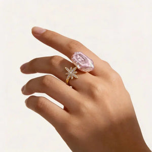Hand wearing a gold ring with a large iridescent pearl and a star-shaped crystal accent