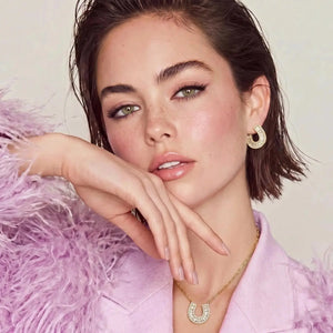 Woman wearing gold hoop earrings and a necklace, sitting on a pink fluffy surface.