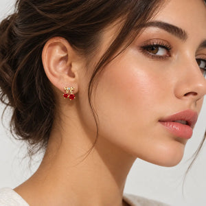 A close-up of a woman's ear wearing a small gold stud earring with three red gemstones, against a clean light gray background.