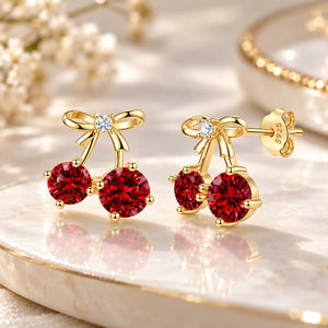 A pair of gold cherry stud earrings with deep red gemstone cherries, bow details, and a small clear center stone, displayed on a polished cream marble surface with soft floral props.