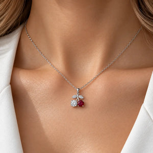 A close-up of a woman's neck and chest wearing a delicate silver chain necklace with a cherry pendant. The pendant features a round red gemstone and a smaller clear gemstone surrounded by a silver leaf design.