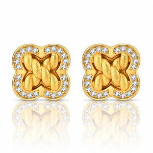 A pair of gold-toned stud earrings with a four-leaf clover shape, featuring a twisted knot design in the center and a border of small clear stones, shown on a white background with a soft reflection below.