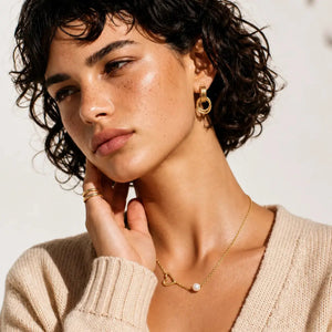 A close-up lifestyle shot of a woman with short curly dark hair wearing gold jewelry, including a gold hoop earring, a delicate gold necklace with a small pearl, and stacked gold rings on her finger, paired with a beige knit sweater.