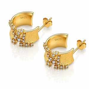 A pair of gold geometric stud earrings with clear gemstones, displayed on a clean white background with a soft reflection below.
