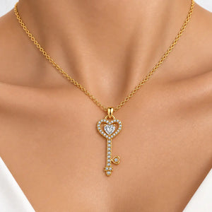 A gold key-shaped pendant necklace with a heart outline and clear stones, worn on a model's neck.
