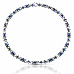 A silver-toned tennis necklace featuring rectangular and round-cut deep blue gemstones, displayed on a clean white background with a soft reflection below.
