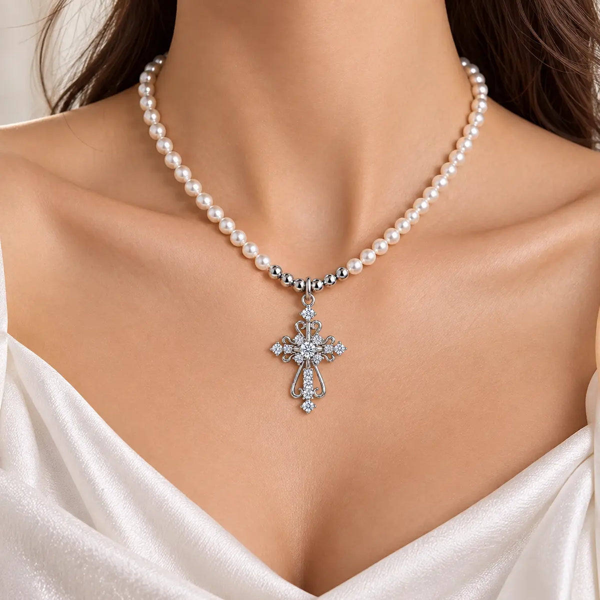 A close-up of a woman's neck and upper chest wearing a pearl necklace with a silver-toned cross pendant adorned with clear stones, paired with a white satin top.