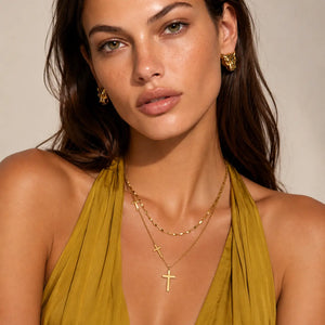 A close-up of a woman wearing gold cross pendants on layered necklaces and matching gold hoop earrings, paired with a mustard yellow halter top against a neutral beige background.