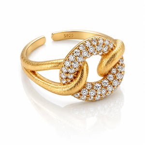A gold adjustable ring featuring a knot-like design with pavé-set clear stones on one side and a textured finish on the other, shown on a white reflective background.