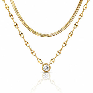 A gold necklace set featuring a double-layered chain design: a thinner snake chain and a thicker oval-link chain with a round bezel-set clear gemstone pendant, displayed on a white background with a soft reflection.