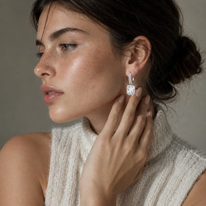 A close-up lifestyle shot of a woman wearing a silver huggie hoop earring with a cushion-cut clear gemstone. She is wearing a cream-colored knit sleeveless top against a neutral gray background.