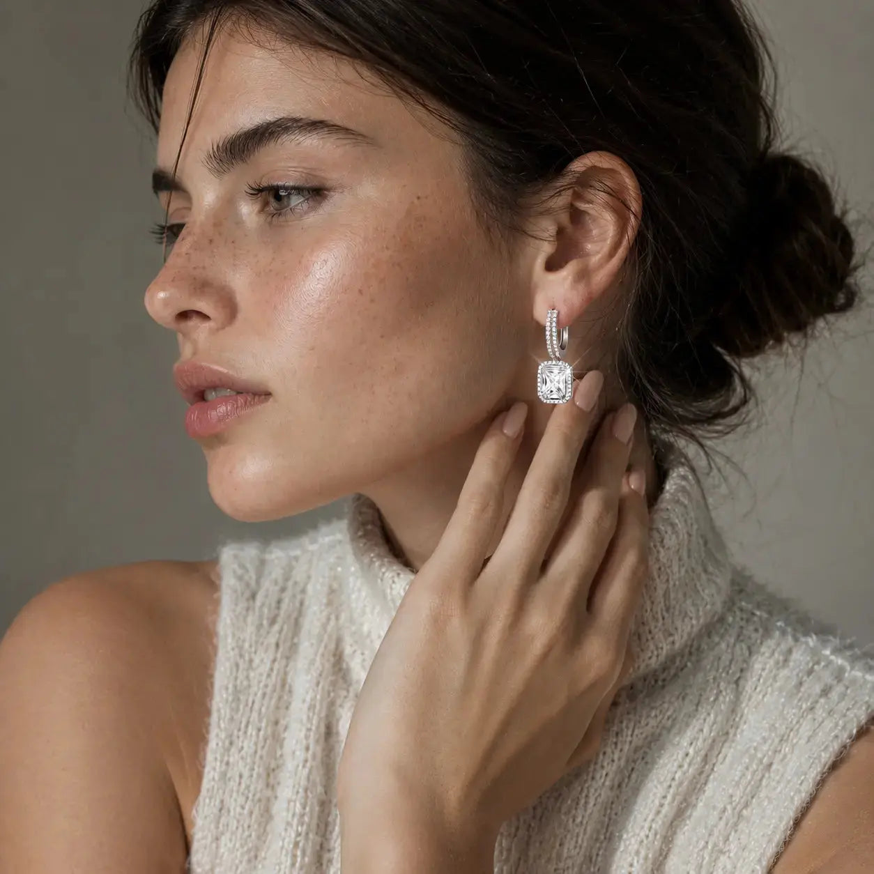 A close-up lifestyle shot of a woman wearing a silver huggie hoop earring with a cushion-cut clear gemstone. She is wearing a cream-colored knit sleeveless top against a neutral gray background.