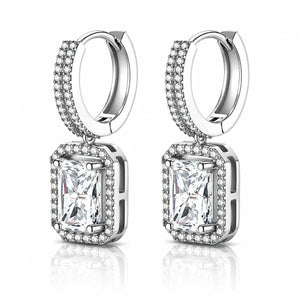 A pair of silver-toned huggie hoop earrings with rectangular emerald-cut clear gemstones surrounded by small pavé-set stones, shown on a clean white background with a soft reflection below.