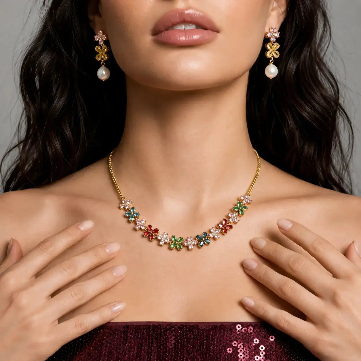 A close-up lifestyle shot of a model wearing a gold floral necklace and matching drop earrings. The necklace and earrings feature gold flower-shaped settings adorned with small multicolored gemstones (pink, green, blue, red, and white) and a single white pearl hanging below each flower.