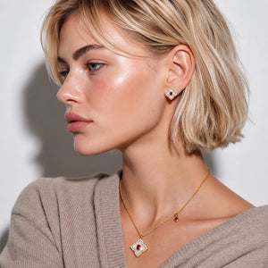 A close-up side profile of a blonde woman wearing a delicate gold necklace with a red and white clover pendant and matching stud earrings, paired with a beige knit sweater against a neutral light gray background.