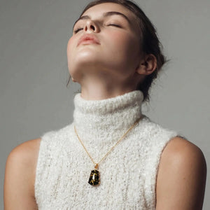 Woman wearing a white turtleneck sweater and gold necklace with a dark pendant against a gray background