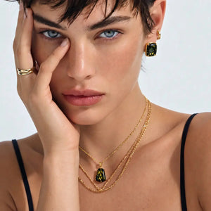 A close-up lifestyle shot of a model wearing a gold layered necklace set with green rectangular gemstone pendants and matching drop earrings.