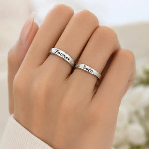 A close-up of a hand wearing two slim silver rings engraved with the words 'Forever' and 'Love' in black script, set against a soft, blurred light background.