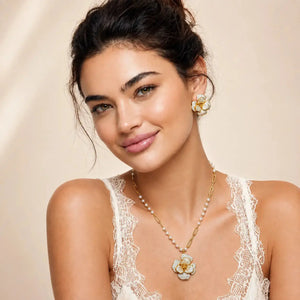 A woman wearing a gold and white flower pendant necklace with a matching flower stud earring, styled with a cream lace camisole against a soft beige background.
