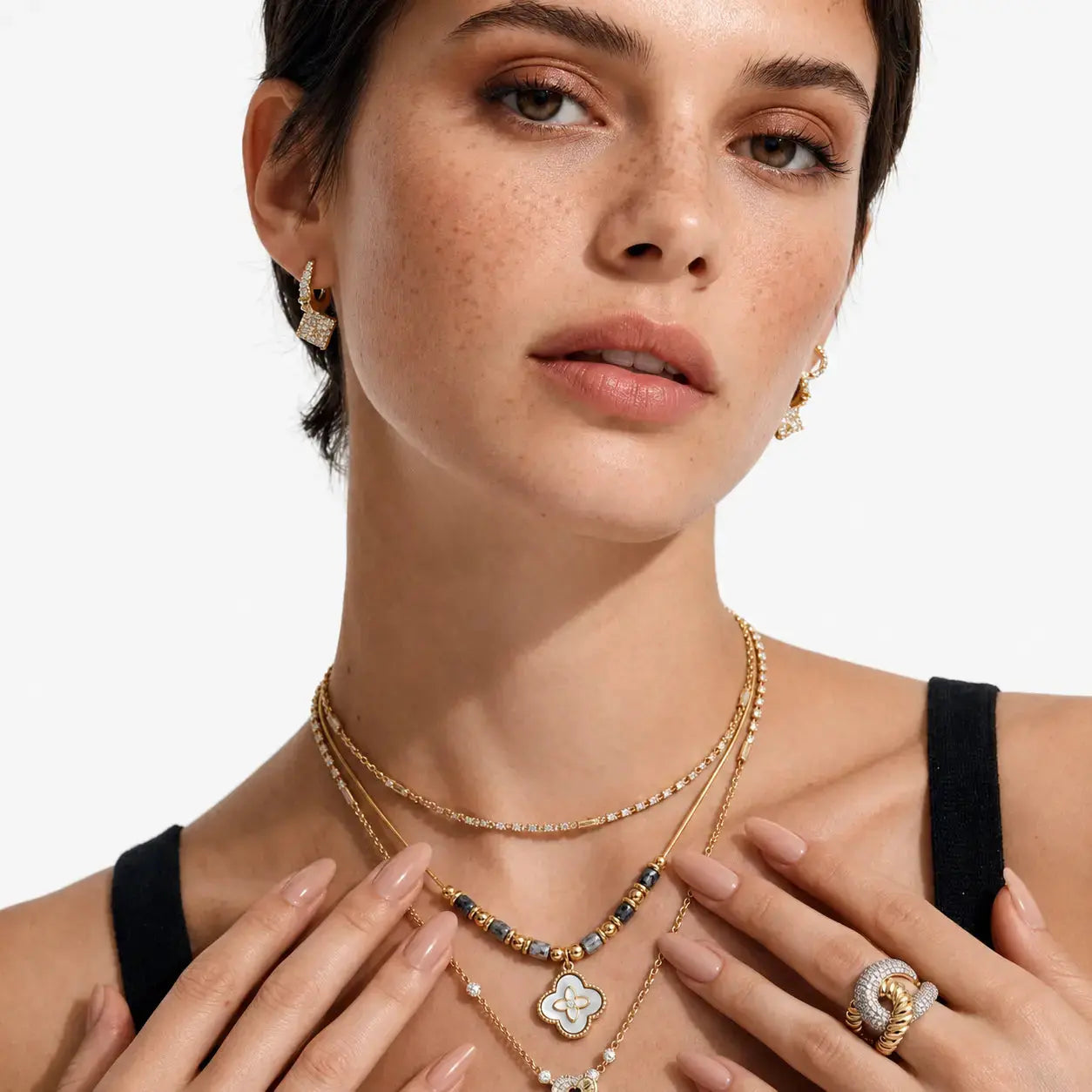 A close-up of a model wearing layered gold necklaces, a beaded charm necklace, a silver and gold ring, and gold hoop earrings against a clean white background.