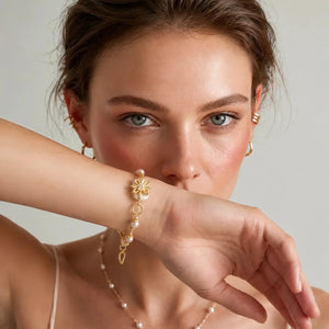 A close-up lifestyle shot of a model wearing a delicate gold chain bracelet with a flower charm and pearl accents on her wrist, paired with matching pearl and gold earrings.