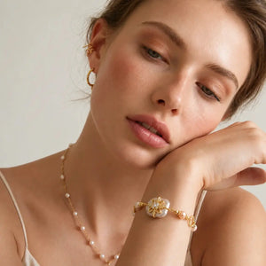 A close-up lifestyle shot of a woman wearing a delicate gold bracelet with a pearl flower centerpiece on her wrist, along with a pearl and gold chain necklace.