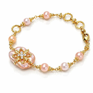 A gold chain bracelet featuring pink freshwater pearls and a large baroque pearl centerpiece decorated with a gold floral brooch and small clear crystals, shown on a white background.